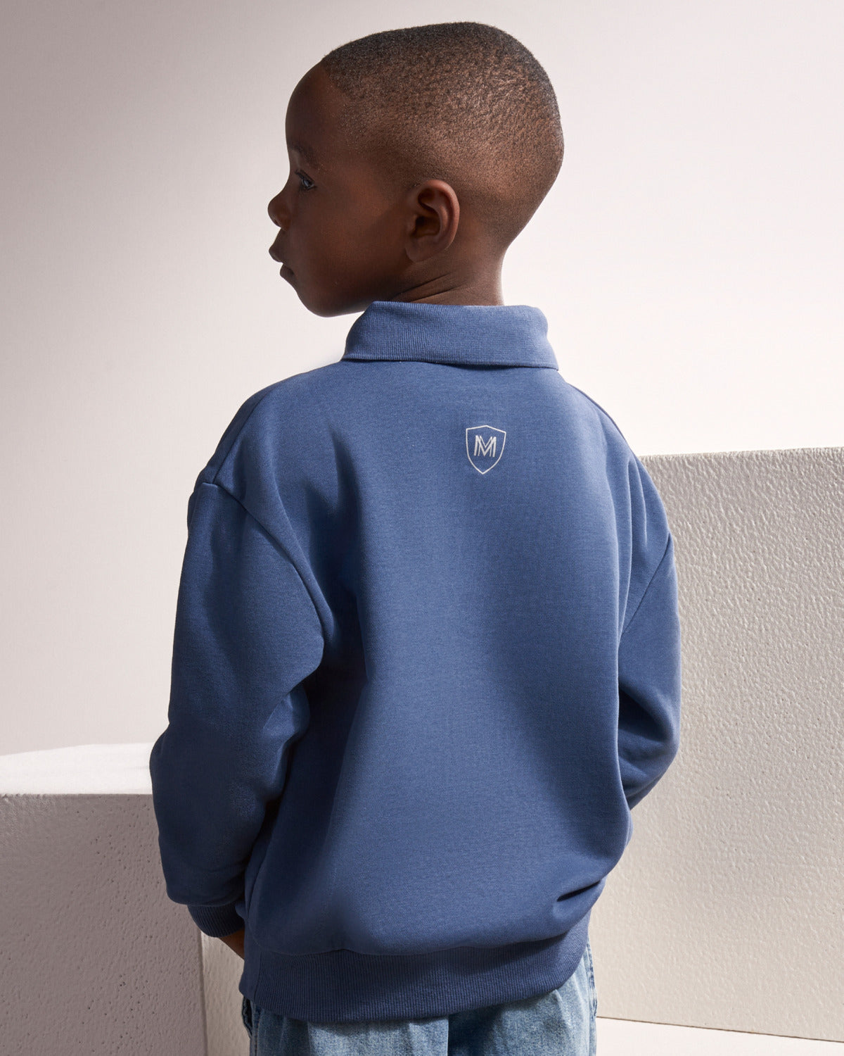 A young boy wearing a blue quarter-zip sweatshirt, shown from the back. The sweatshirt features a small white shield logo with an 'M' on the upper back. The boy is standing against a minimalist, light-colored textured wall.