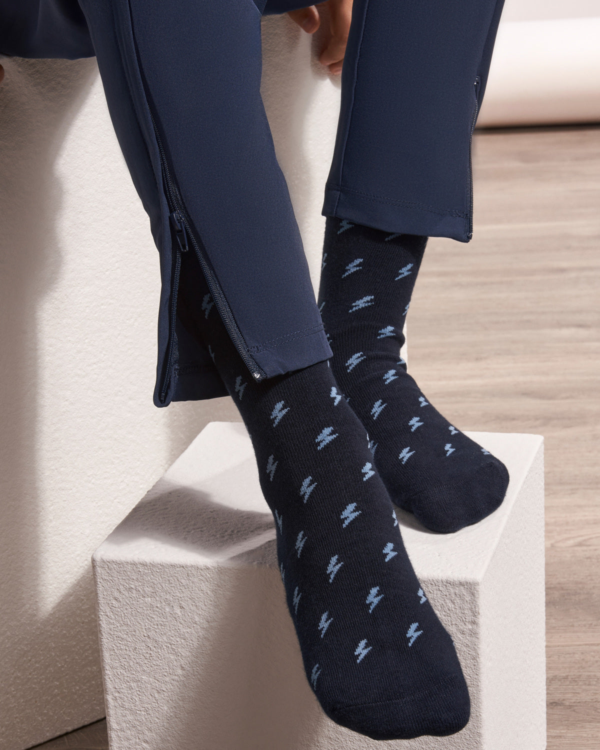 A close-up of a person wearing navy blue patterned crew socks with light blue lightning-bolt motifs, paired with navy pants, sitting on a white block.