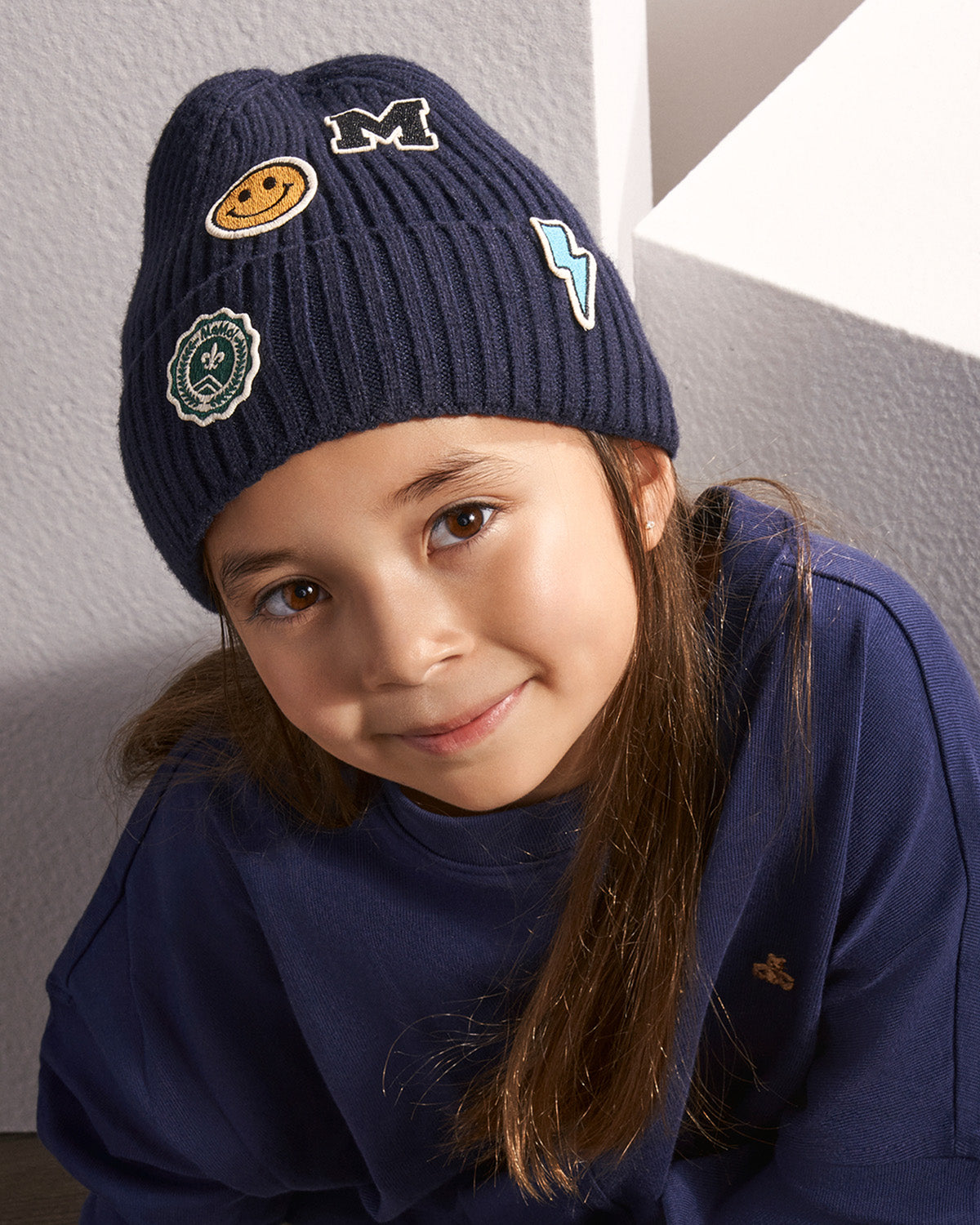 A young girl with long brown hair wearing a navy blue ribbed beanie with embroidered patches and a matching navy blue sweatshirt, posing against a textured gray wall.