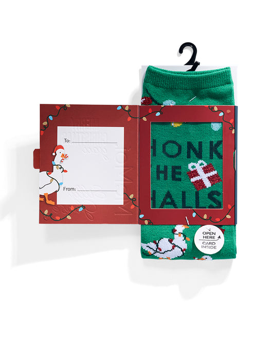 A Christmas-themed sock advent box on a white background. The red cardboard box has holiday decorations, a chicken illustration, and a writing panel with 'To' and 'From.' fields. A green sock with 'HONK THE HALLS' and a gift graphic is partially pulled out, with additional green socks visible below and a black hanger at the top.