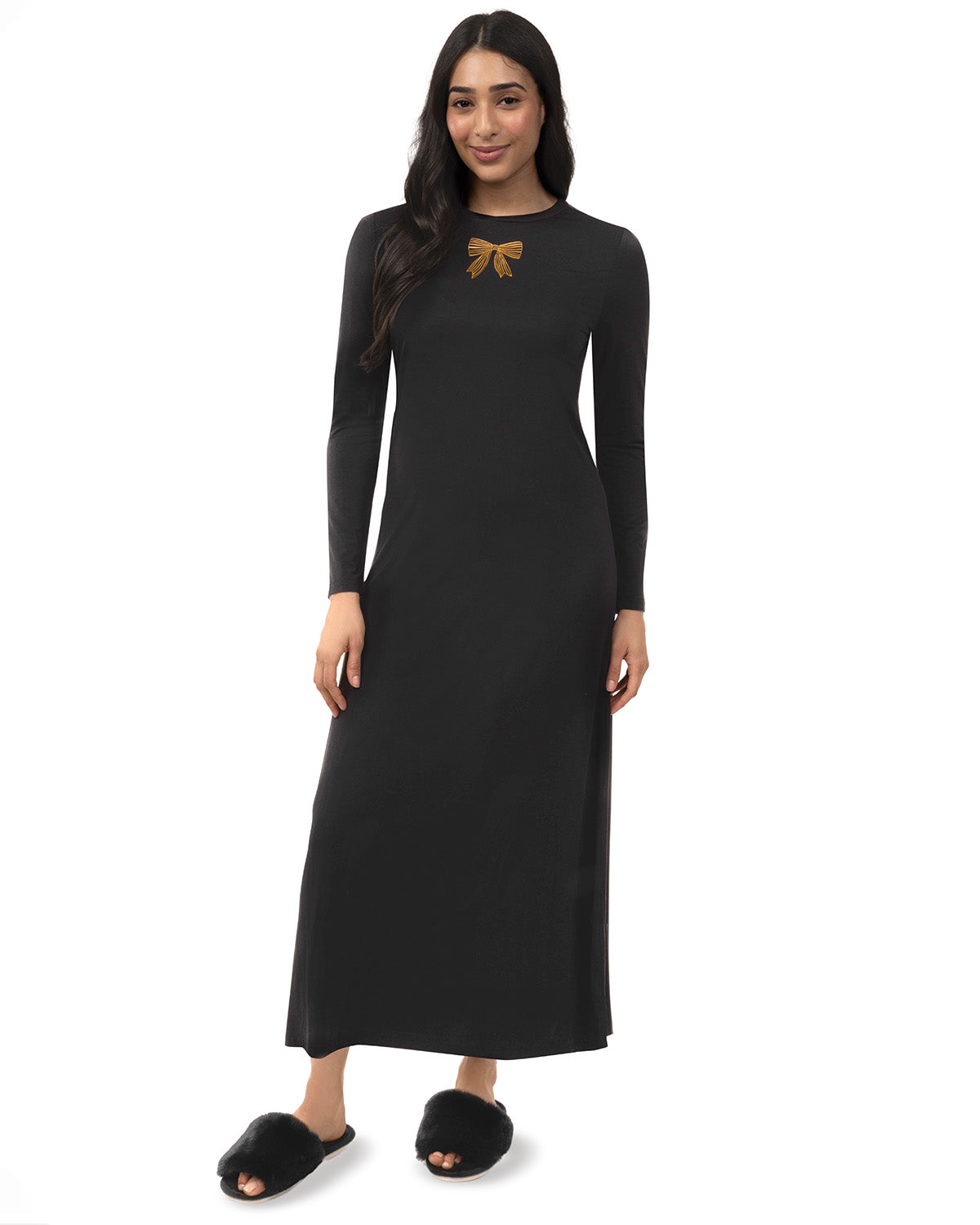 A woman modeling a long-sleeve black maxi dress with a small gold bow detail on the chest, paired with black fuzzy slippers against a plain white background.