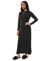 A woman modeling a long-sleeve black maxi dress with a small gold bow detail on the chest, paired with black slippers, against a plain white background.