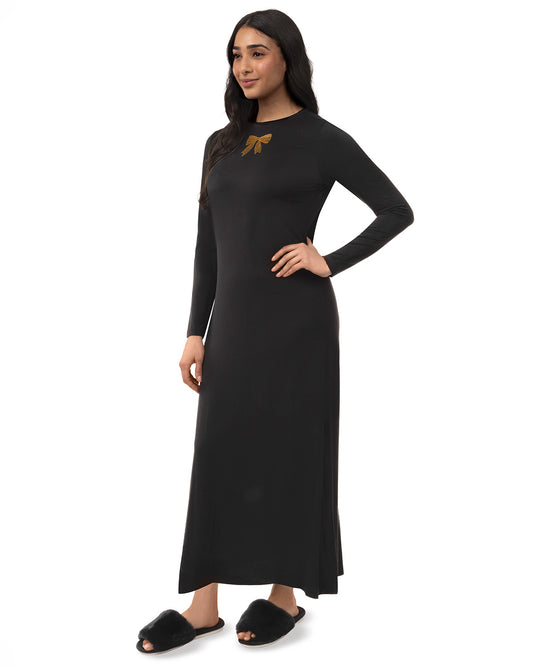 A woman modeling a long-sleeve black maxi dress with a small gold bow detail on the chest, paired with black slippers, against a plain white background.