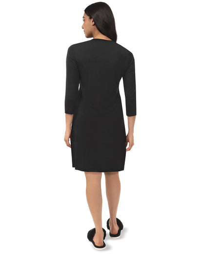 A woman shown from the back wearing a black knee-length dress with three-quarter sleeves, paired with black slip-on shoes, against a plain white background.
