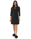A woman modeling a black three-quarter sleeve shift dress with a small gold bow detail on the chest, paired with black fuzzy slippers against a plain white background.