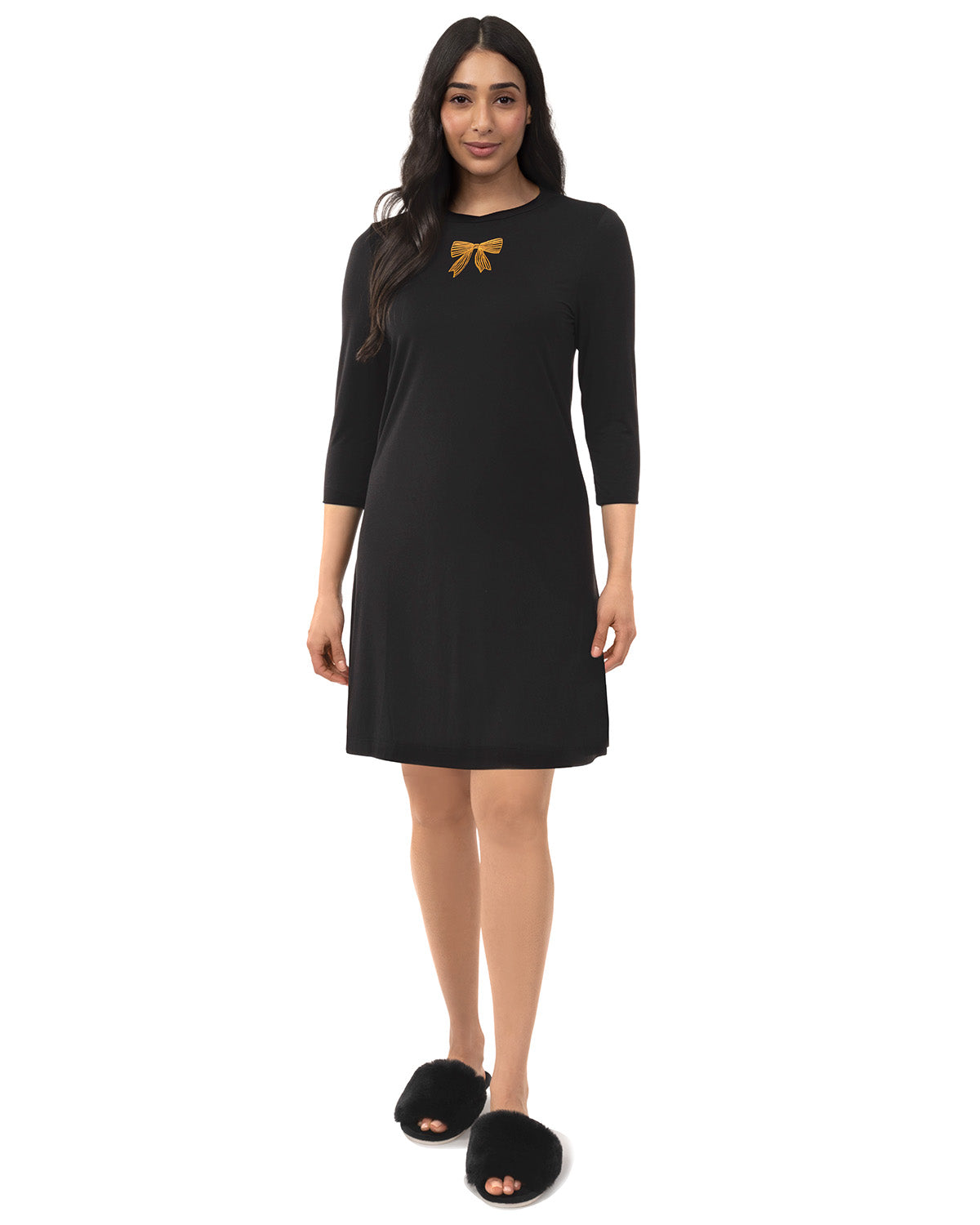 A woman modeling a black three-quarter sleeve shift dress with a small gold bow detail on the chest, paired with black fuzzy slippers against a plain white background.