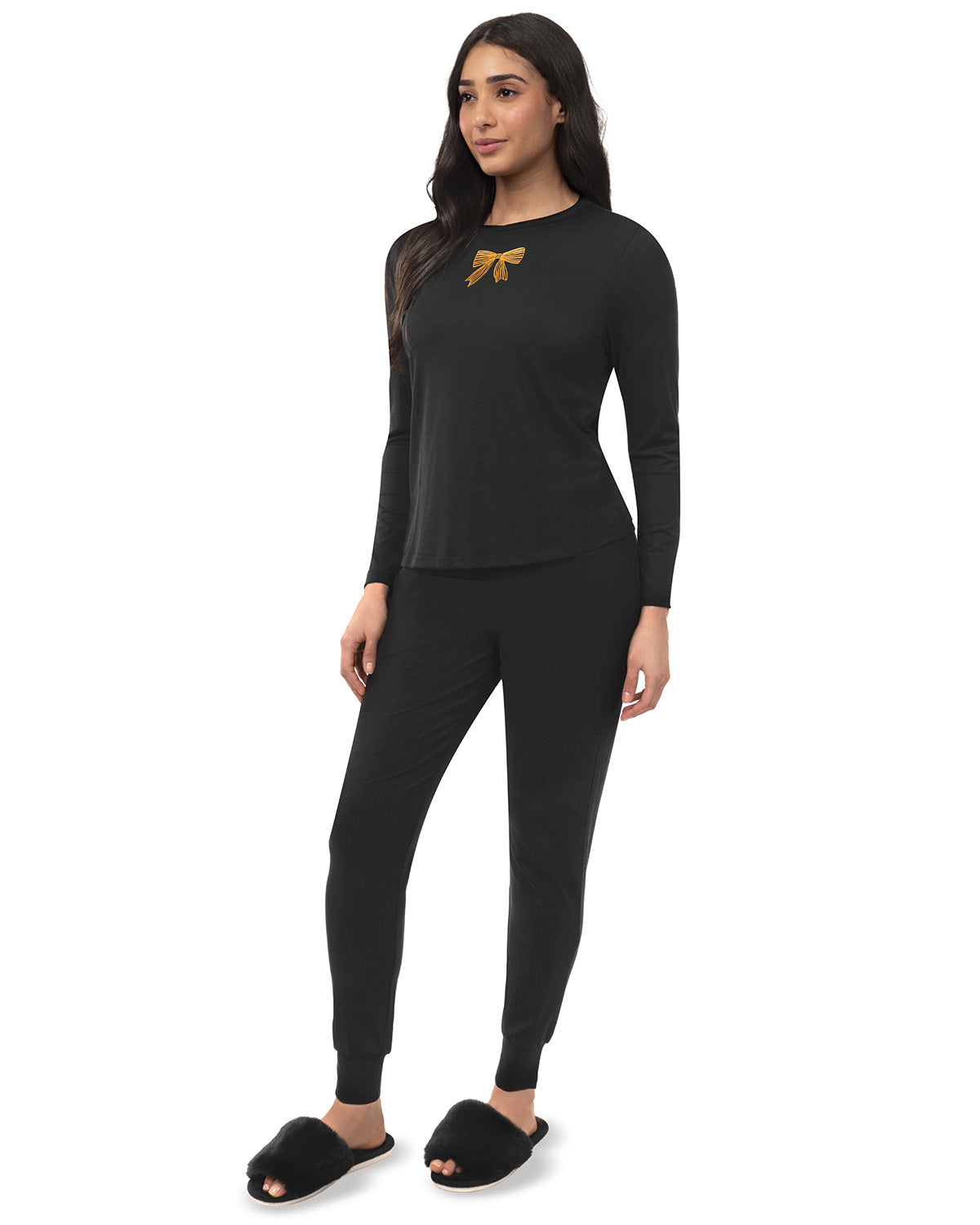A woman modeling a black two-piece lounge set consisting of a long-sleeve top with a small gold bow graphic on the chest and matching black jogger pants, paired with black slippers against a plain white background.