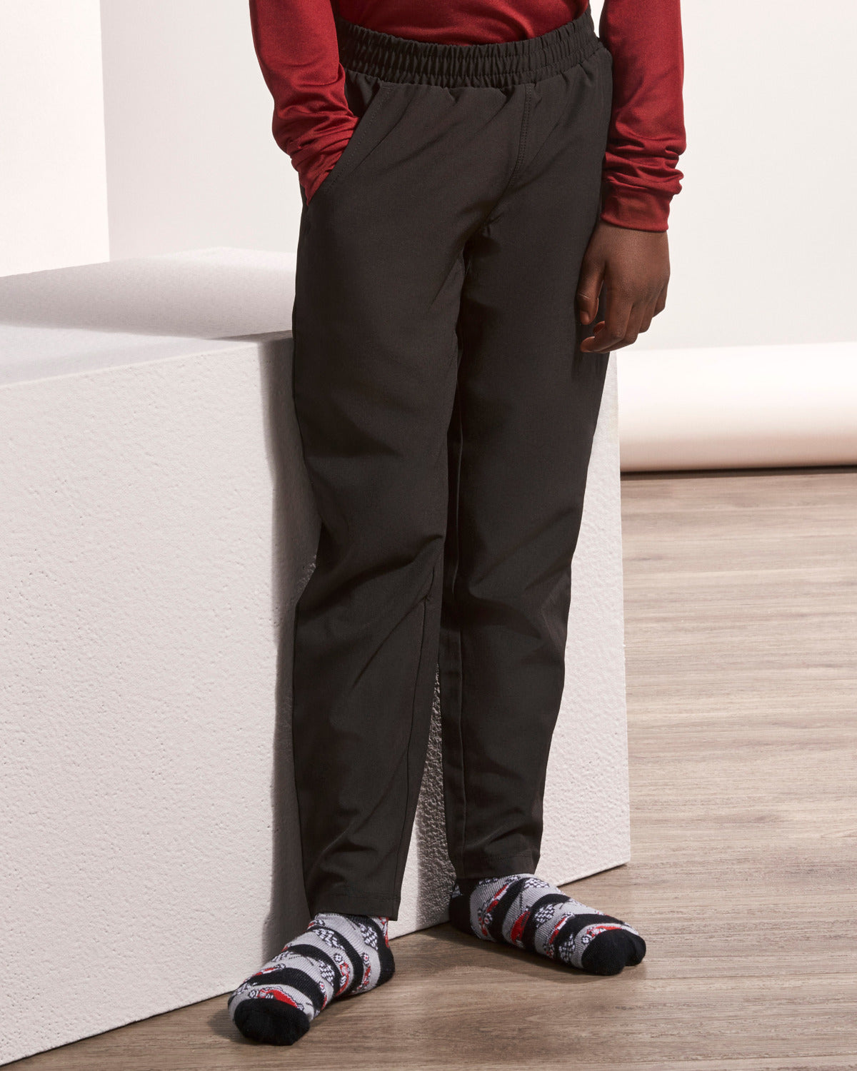A person wearing dark charcoal gray jogger-style pants with an elastic waistband, paired with a red long-sleeve top and patterned socks, standing next to a white block on a light wood floor.