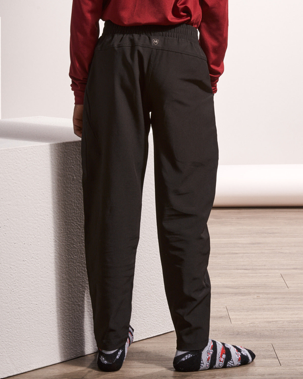 A person shown from the back wearing loose black pants, a dark red long-sleeve top, and patterned socks, standing indoors against a white wall and light wood floor.