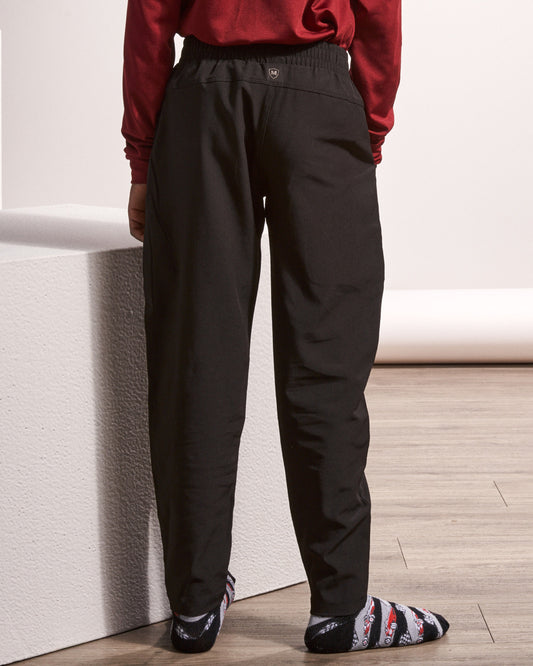 A person shown from the back wearing loose black pants, a dark red long-sleeve top, and patterned socks, standing indoors against a white wall and light wood floor.