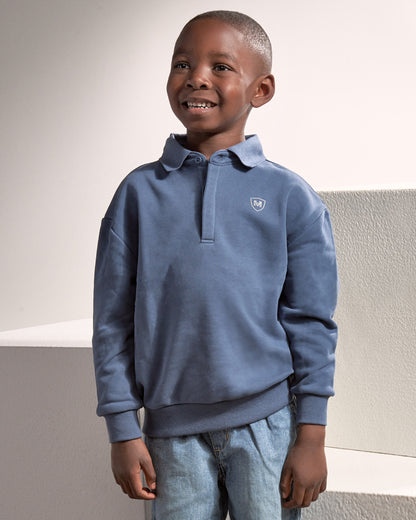 A young boy smiling and looking to the side, wearing a dusty blue long-sleeve polo sweatshirt with a small shield logo on the left chest, paired with light blue jeans.