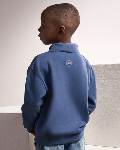 A young boy wearing a blue quarter-zip sweatshirt, shown from the back. The sweatshirt features a small white shield logo with an 'M' on the upper back. The boy is standing against a minimalist, light-colored textured wall.