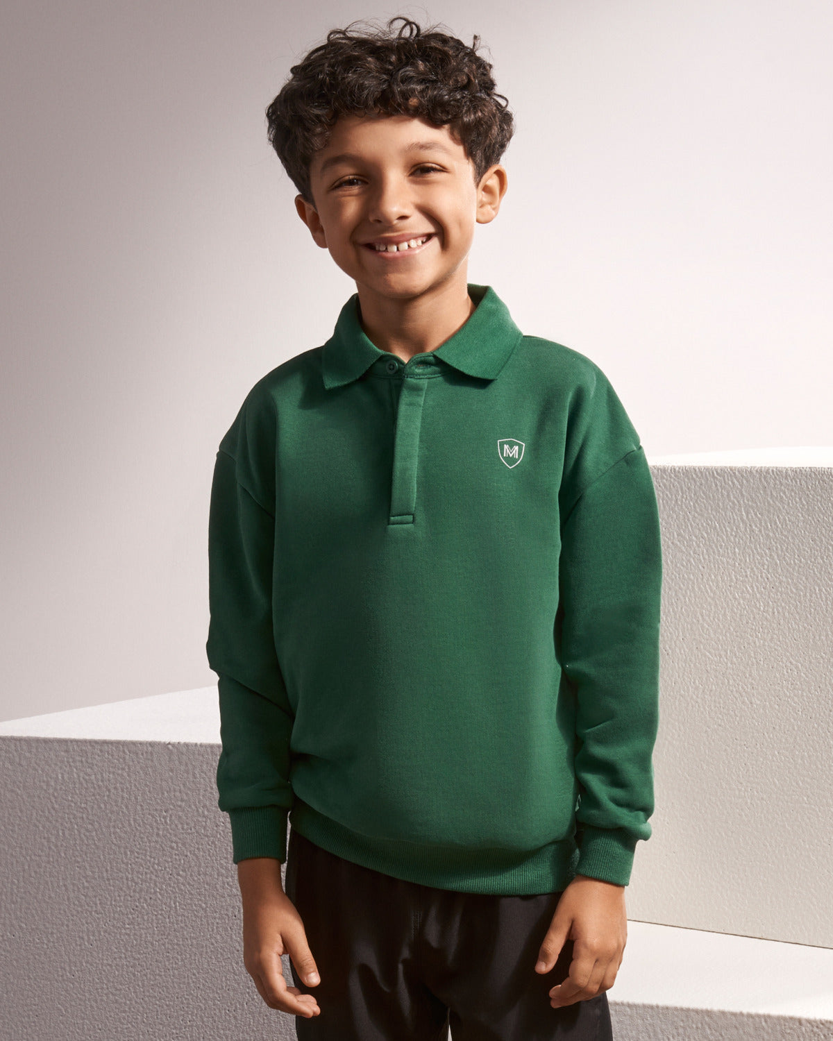 A young boy smiling while wearing a dark green long-sleeve polo shirt with a small shield logo on the left chest, paired with black pants, set against a light gray textured background.