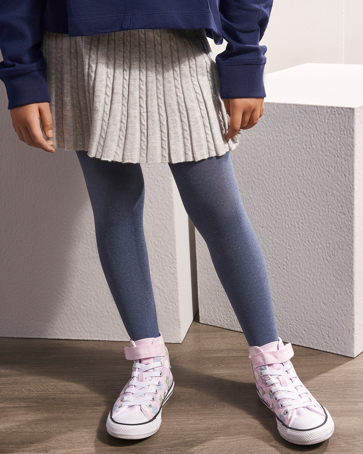 A child wearing a gray pleated cable-knit mini skirt, slate blue tights, and light pink high-top sneakers, standing in front of textured white blocks.
