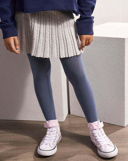 A child wearing a gray pleated cable-knit mini skirt, slate blue tights, and light pink high-top sneakers, standing in front of textured white blocks.