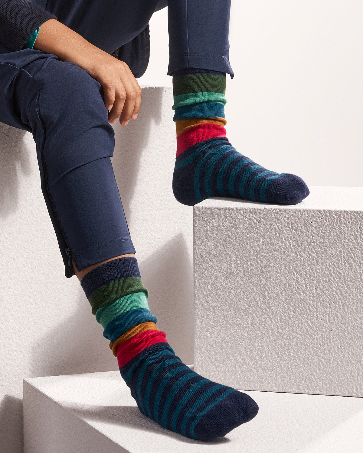 A person seated on white geometric steps wearing navy pants and colorful striped crew socks. The socks feature horizontal bands of dark teal, navy, red, orange, green, and teal.