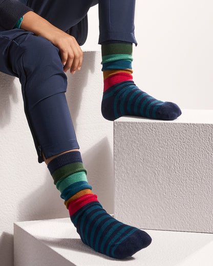 A person seated on white geometric steps wearing navy pants and colorful striped crew socks. The socks feature horizontal bands of dark teal, navy, red, orange, green, and teal.