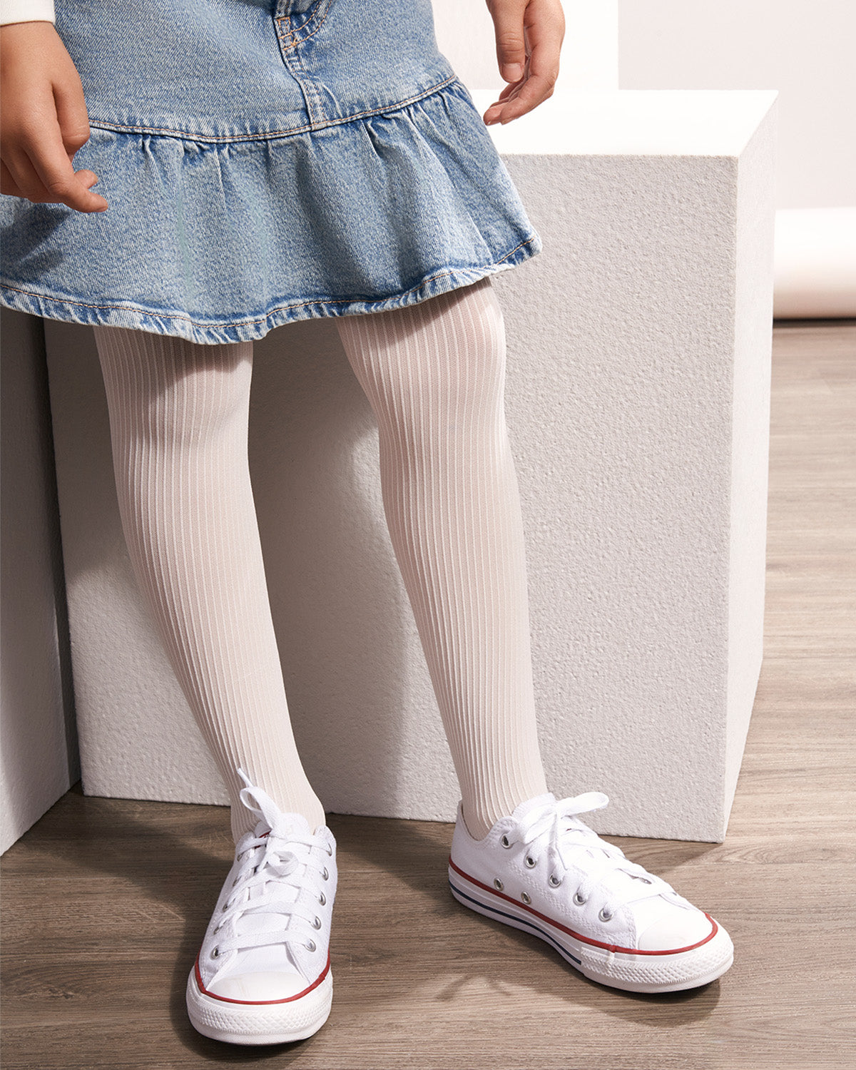 A child wearing light beige ribbed tights with white low-top sneakers and a light blue denim ruffled skirt, standing next to a white block on a wood floor.