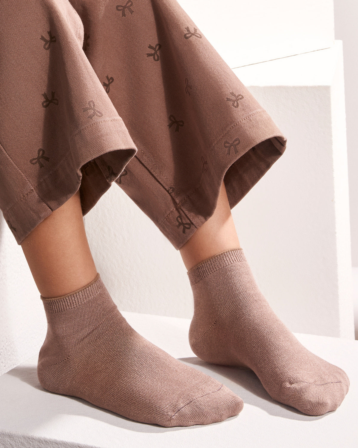 A close-up of a person's feet wearing taupe-brown ankle socks with a matching patterned trouser leg, sitting on a white step.