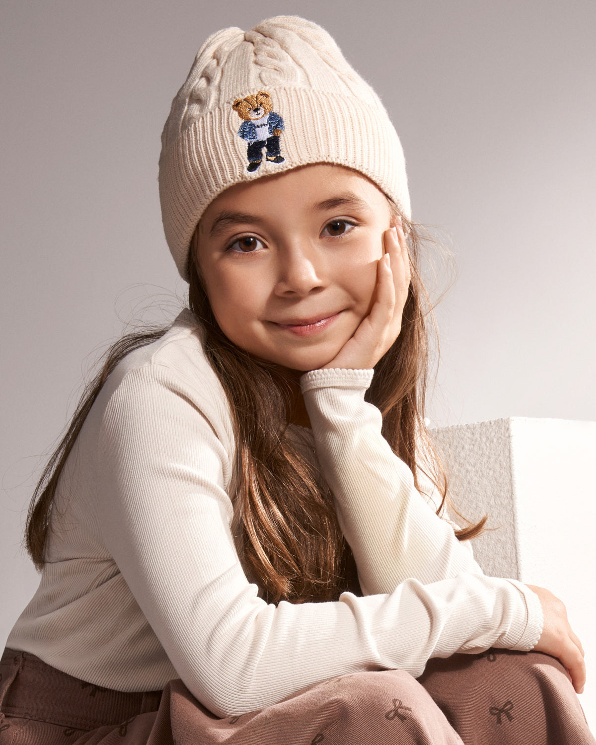 A young girl with long brown hair sits smiling, wearing a cream cable-knit beanie with an embroidered teddy bear logo, a cream long-sleeve top, and brown pants against a neutral gray background.