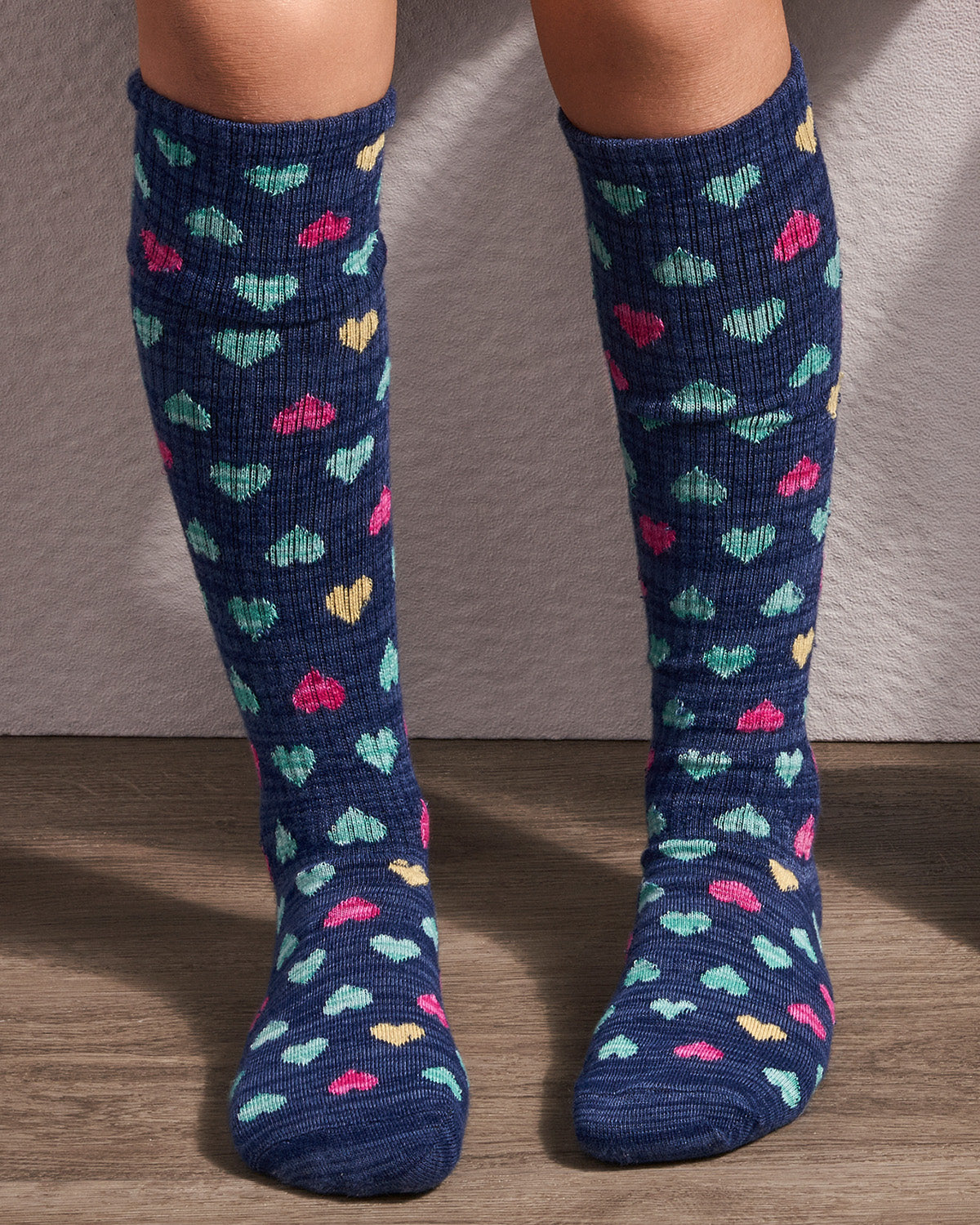 A pair of navy blue knee-high socks with a colorful knit heart pattern, worn on a person's legs against a neutral wall and wood floor.