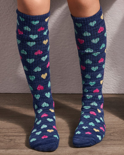 A pair of navy blue knee-high socks with a colorful knit heart pattern, worn on a person's legs against a neutral wall and wood floor.