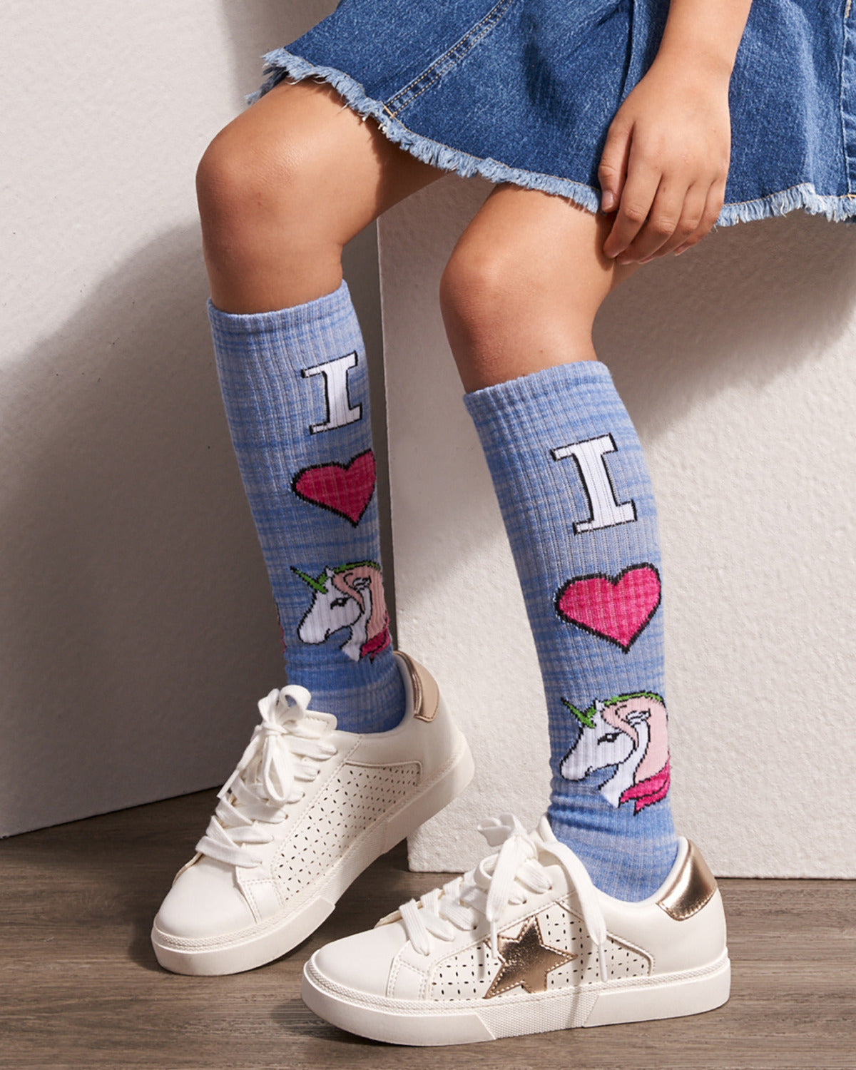 A person seated against a light wall wearing light blue knee-high socks with a plaid pattern, large white letters, pink hearts, and unicorn illustrations, paired with white sneakers.