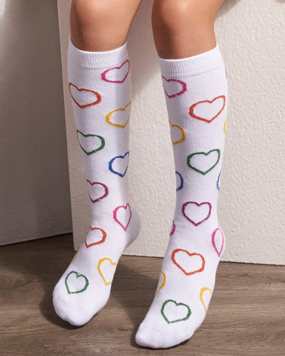 White knee-high socks with a colorful pattern of outlined hearts in pink, orange, green, blue, and yellow, worn on a person's legs against a neutral wall and wood floor.