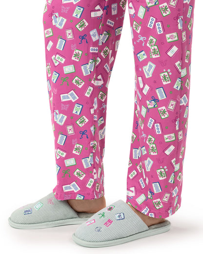 Pink patterned pajama pants with a playful print of postcards, money, and small icons, worn with light gray slip-on slippers on a white background.